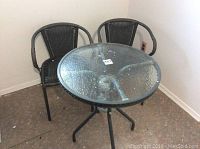 Photo showing round glass-top garden table and two black wicker chairs against wall
