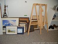 Wooden folding art easel and assortment of framed pictures and paintings arranged against a wall on carpeted floor.