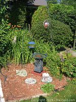 Outdoor garden view showing bird bath on pedestal surrounded by plants and two shepherd's hooks with decorative hanging items.