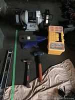 Photo showing Delta bench grinder, blue and black anvil, two hammers, and a toolbox with wrenches and sockets on the floor.