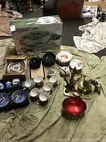 Wide view of the lot showing Paderno boxed pans, various Asian-themed ceramic plates, bowls, cups, brass decanter and tray, decorative lamp button.