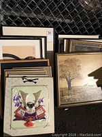 Multiple framed artworks stacked leaning against a chain link fence, showing different styles and themes. Decorative samurai helmet artwork visible in front.