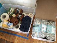 Plastic bin with multiple skeins and cones of yarn in various colors including yellow, brown clove, sage green and ecru cones plus variegated Bernat skeins in box beside bin.