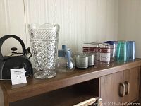 Photo of the black and silver microwaveable 3-cup tea kettle, large clear crystal vase with diamond pattern, assorted clear glassware, Coca-Cola, Scooby Doo, and Tervis tumblers on wooden shelf