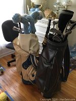 Two golf bags side by side on a hardwood floor near a chair and table, showing blue knit headcovers on some clubs in the beige bag and various clubs in the black bag.
