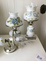 Three lamps grouped on a white surface: two white milk glass table lamps with blue rose hand painted shades and bases, the tallest lamp showing a noticeable chip on the ruffled top shade. A wall lamp with a missing top shade is visible behind.