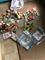 Photo showing over 20 skeins of assorted colored yarn, multiple needlepoint canvases with finished floral designs, some framed or wrapped in plastic.