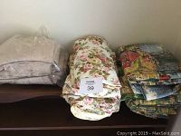 Three fabric piles displayed on wooden surface: cream lining in plastic, floral fabric with roses, and scenic fabric with houses.