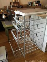 White metal closet organizer with four wire baskets of varying sizes placed in a room with wooden floors and furniture around.