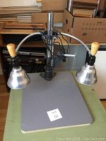 Full view of the photographic stand with two adjustable lights, showing the gray base, vertical metal pole, two metal light shades with wooden handles, and adjustable arms.