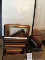 Photo shows a variety of mostly wooden and some metal picture frames with glass, stacked and standing in boxes and leaning against a wall.