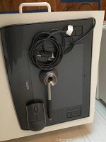 Top view of Wacom Intuos 3 tablet in dark gray with pen placed in pen holder stand, USB cord coiled on tablet, and mouse placed on the tablet.