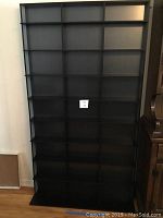 Full front view of the black media storage shelf showing 9 shelves arranged in 3 columns, placed against the wall.
