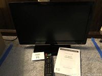 Front view of Proscan 19 inch LED TV, power indicator on, remote control placed in front, operating instructions manual visible beside it.