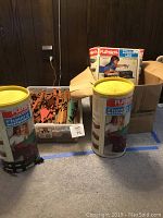 Photo showing two PlaySkool Lincoln Logs cylinders, a clear bin full of wooden Lincoln logs, and a cardboard box filled with additional logs and roof/window pieces.