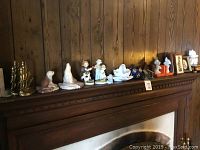 Wide view of assorted knickknacks on a mantle including figurines of seals, Hummel and Holly Hobby style children, milk glass swan, and framed pictures.