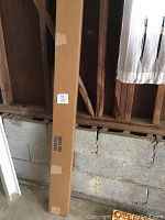 Photo of the unopened, long rectangular brown cardboard box labeled 'TWIN/FULL BED FRAME' leaning against a wall in a room with exposed wooden beams and cinder block wall.