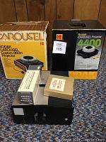 Two Kodak Carousel slide projectors with original boxes and additional smaller boxes, arranged on carpeted floor in front of wood panel wall.