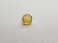 Round cut faceted Ethiopian opal gemstone resting on white surface showing yellow body color and rolling green flash effect.