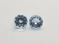 Two round diamond cut light blue topaz stones displayed side by side on white background, showing facets reflecting light.