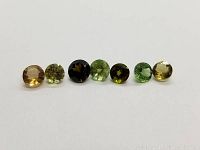 Seven round cut multicolor tourmaline gemstones displayed in a single row on a white background. Colors include yellow, green, dark green, and near black shades. The stones have a diamond cut faceting style.