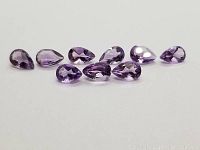 Clear image showing nine pear cut bright purple amethysts laid out in a row on a white surface.