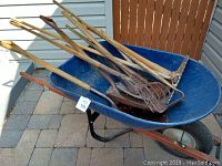 Blue wheelbarrow holding various garden tools including shovels, rakes, pitchfork, hoe, pick, sledge hammer, and axe.