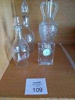 Photo showing three clear glass decanters and a Waterford crystal clock with a small round clock face on the front. The decanters include one tall with two handles and etched pattern, and two others with glass stoppers.