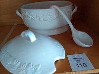 Photo of white ceramic soup tureen with embossed fruit and vegetable designs, matching cover, and ladle.