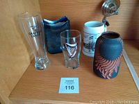 Photo showing two pottery containers, one brown with carved design and one blue with wavy top, alongside a cream-colored Curacao stein with metal lid and two beer glasses, one tall with text and one shorter with a harp emblem.