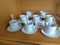 Eight white espresso cups on matching saucers, with three white pottery spoons inside some cups, displayed on wooden shelf.