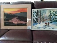 Photo showing both framed paintings on a brown leather couch: the watercolor of a river at sunrise on the left and the acrylic scene of two hunters in snow on the right.