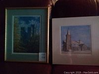 Two framed cathedral and church prints placed side by side on a dark chair. The cathedral print is framed with green matting and gold frame; church print is in white frame and white matting.