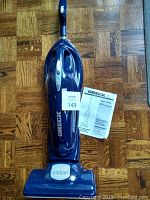 Full view of blue Oreck XL cordless electric broom on wooden floor, with user guide beside it.