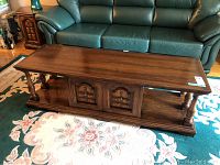 Front view of rectangular wooden coffee table with two-door central cabinet and turned wood pillars on each side, resting on floral carpet near green leather couch and wooden side table.