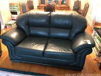 Green leather love seat shown from front angle, placed in a room with wooden flooring and dining table in background.
