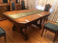 Wooden dining table seen from above showing a green-beige fabric runner over its top, four surrounding chairs, and detailed wood grain surface with scratches.