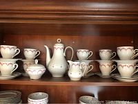 Front view of entire coffee set displayed on wooden shelf showing cups, saucers, coffee pot, sugar bowl, and creamer with pink maroon patterns.