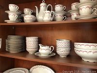 Shelf showing stacked plates on left and multiple teacups with matching saucers on top shelf. Also creamers and gravy dish present in white porcelain with pink rose floral designs and gold trim.