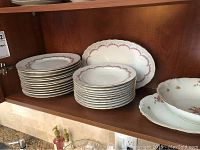 Porcelain kitchenware on wooden shelf, includes stacks of plates, bowls, and serving platters with pink floral and gold trim.