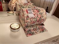 Floral patterned fabric jewelry box closed next to small round ceramic box with gold-rimmed lid on a white surface.