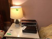 Photo showing the two clock radios and the white ceramic table lamp with beige shade and tassel decorations on a white surface.
