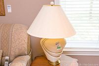Ceramic table lamp shown standing on a wooden side table next to a patterned upholstered sofa, under window light.