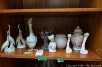 Grouped image showing figurines including two white birds, large white swan with gold beak, iridescent Capodimonte birds on branch, white cat with gold bow, floral lidded jar and a white floral pitcher