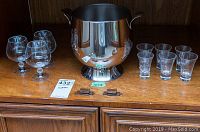 Front view of the snifter glasses, shot glasses and stainless steel ice bucket placed on a wooden shelf.