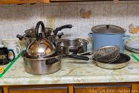Photo showing assorted pots, pans, kettle, strainer basket, with a blue enameled pot and several stainless steel items.