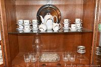 Full view of tea set cups, teapot, creamer, sugar bowl, and glassware displayed in wooden cabinet.