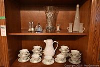 Wooden shelf holding 11 teacups stacked in pairs, 12 saucers, tall clear glass vase, frosted glass dual-handle vase, salt and pepper shakers in metal holder, and thermal coffee pot.