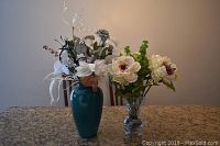 Full view of two arrangements of faux flowers on a granite countertop; one in teal ceramic vase and one in clear pedestal glass vase.