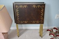 Four vintage folding TV trays standing upright with tops facing forward, showing decorative brown tops with gold floral border pattern and gold metal legs.